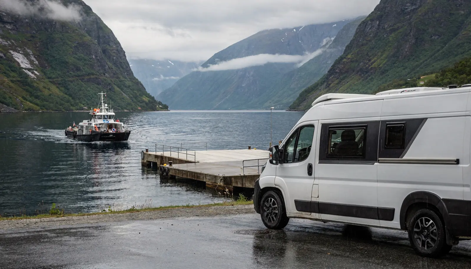 Un camping-car moderne attend à un embarcadère de ferry face aux fjords de Norvège, entouré de montagnes escarpées et d'eau bleu-gris sous un ciel couvert.