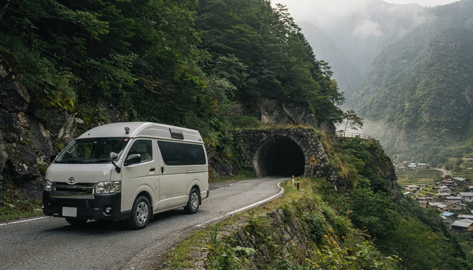 Un camping-car compact circule sur une route sinueuse des Alpes japonaises, entouré de forêts abruptes et de brume, avec l'entrée d'un tunnel en pierre visible devant.