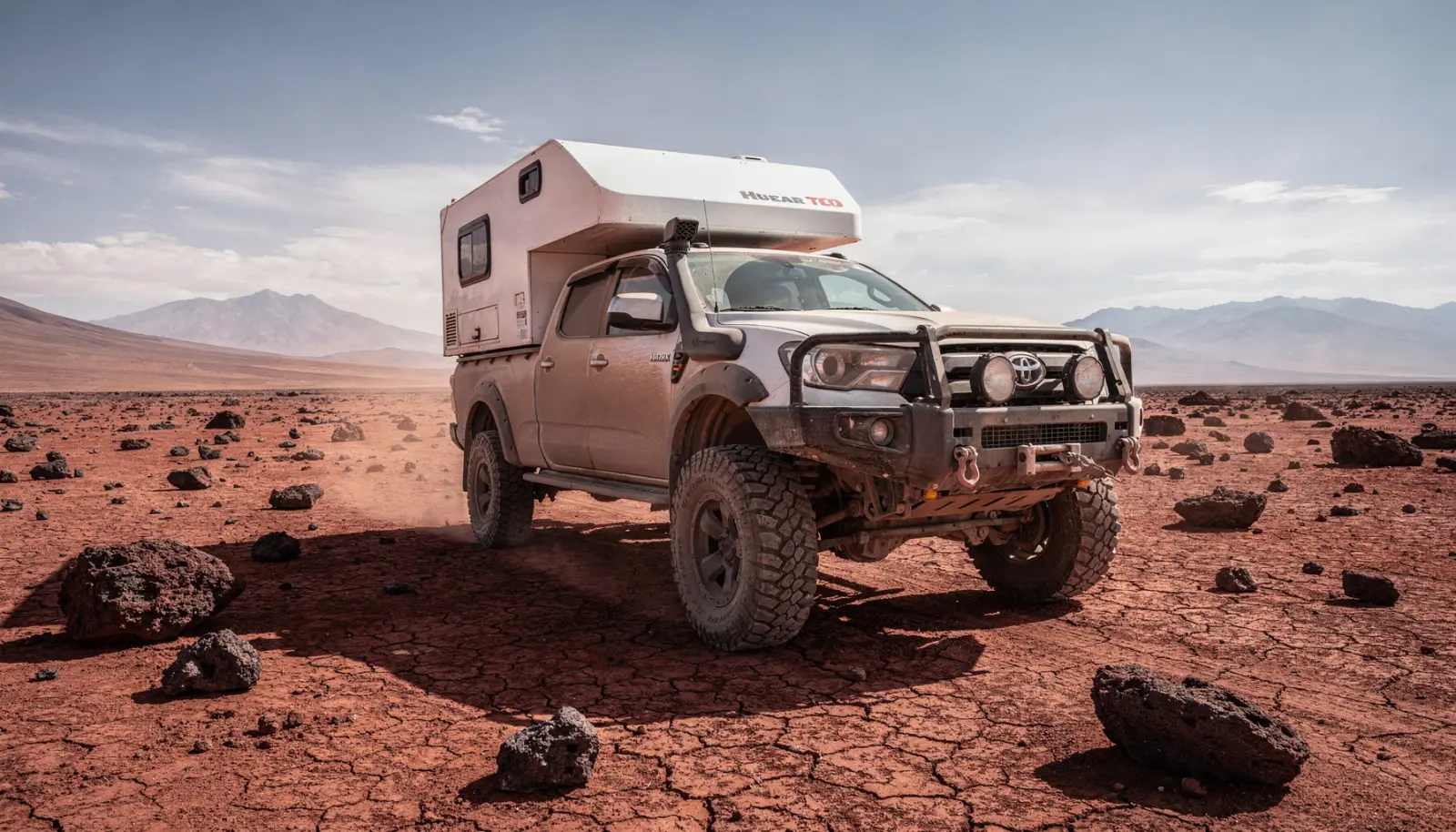 Camping-car 4x4 sur châssis pick-up posé sur une plaine désertique rougeâtre, montagnes arides en arrière-plan