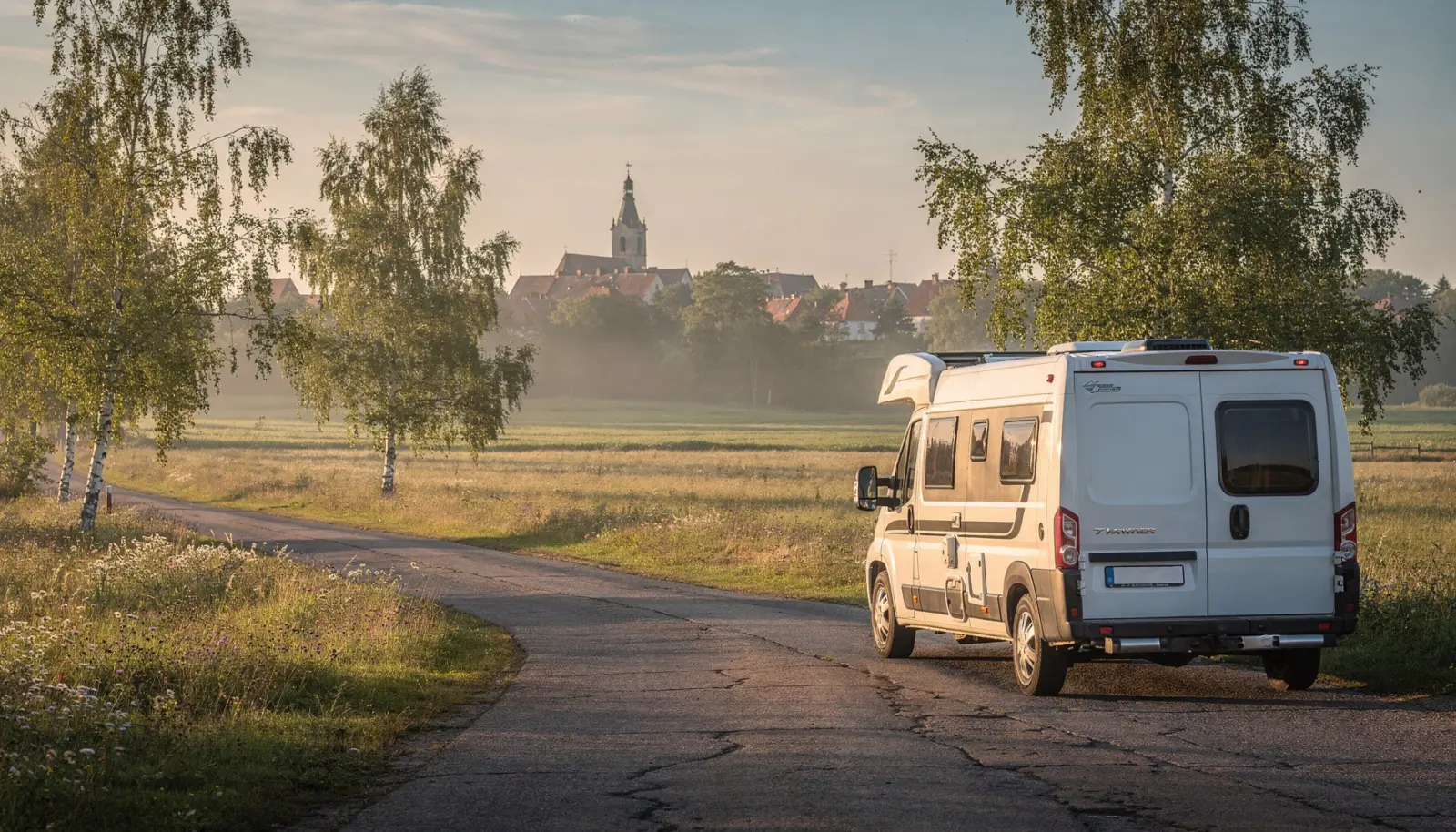 Camping-car garé sur une route de campagne estonienne au lever du soleil, silhouette de vieille ville au loin