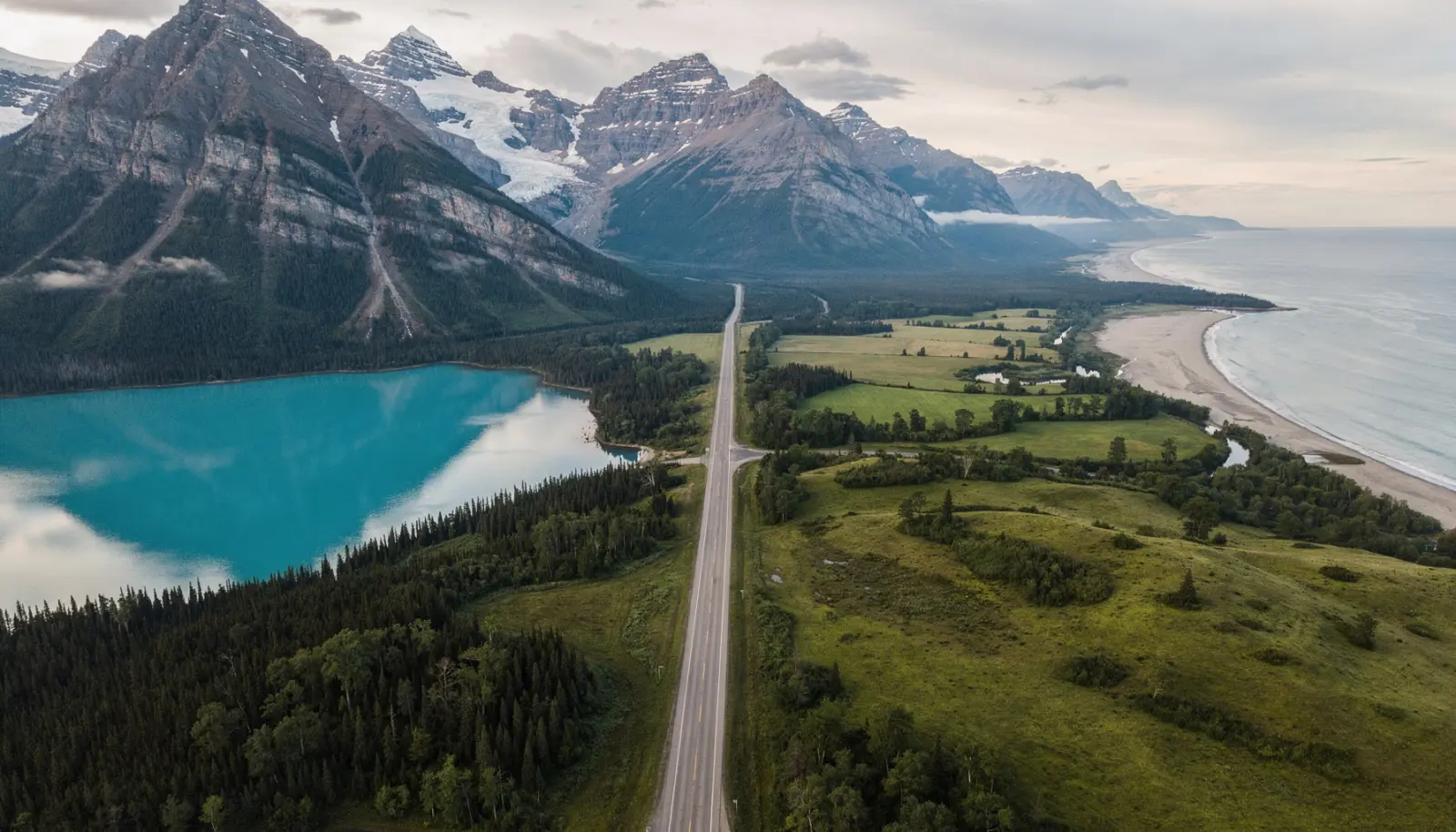 Vue aérienne d'un paysage canadien coupé en deux : montagnes enneigées et lac glaciaire d'un côté, collines verdoyantes et côte de l'autre, reliés par une route